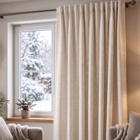 Vorhang aus wollweißem Thermochenille vor Fenster, dichter Faltenwurf, gemütliches Wohnzimmer mit Sessel und Pflanze