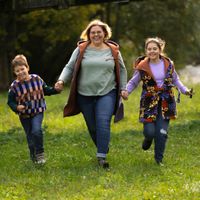 Eine Mama und ihre beiden Kinder rennen über eine Herbstwiese und tragen Jacken und Westen aus Teddyplüsch in orange, lila, grün