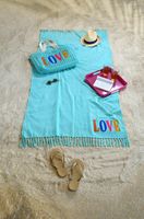 Strandtuch Hamamtuch Plaid Fouta LOVE mit Fransen in der Farbe türkis liegt auf hellem Sand, davor Sandalen, darauf Tasche, sonnenbrille, Strohhut, Tablett, Buch und Gläser mit Cola
