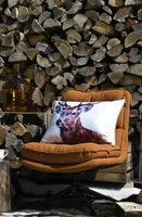 Hirschkissen auf braunem Sessel vor Holzstapel – gemütliche Herbst- und Winterdeko mit Chalet-Charme