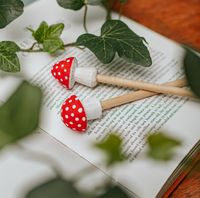 Zwei Holzbleistifte mit Fliegenpilz-Kappen liegen auf einem offenen Buch, umgeben von grünen Efeublättern