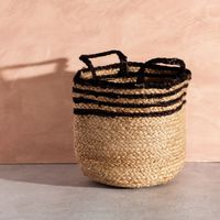 Dekorativer, geflochtener Korb von Sass & Belle aus Naturmaterial. Der runde Korb mit Henkeln ist teilweise schwarz gestreift. Er eignet sich zur Aufbewahrung und Dekoration und ist 28×20 cm groß - vor roséfarbener Wand