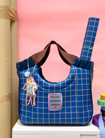 Handtasche aus dunkelblauem Kunstleder mit ockerfarbenem Karomuster, braunen Tragegriffen und dekorativen Anhängern.