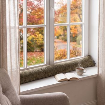 SCHÖNER LEBEN. Zugluftstopper Soft Teddy Deluxe moosgrün für Fenster und Türen, waschbar, verschiedene Größen Zugluftstopper in moosgrün aus softem Stoff liegt auf einer Fensterbank neben einer Tasse und einem aufgeschlagenen Buch