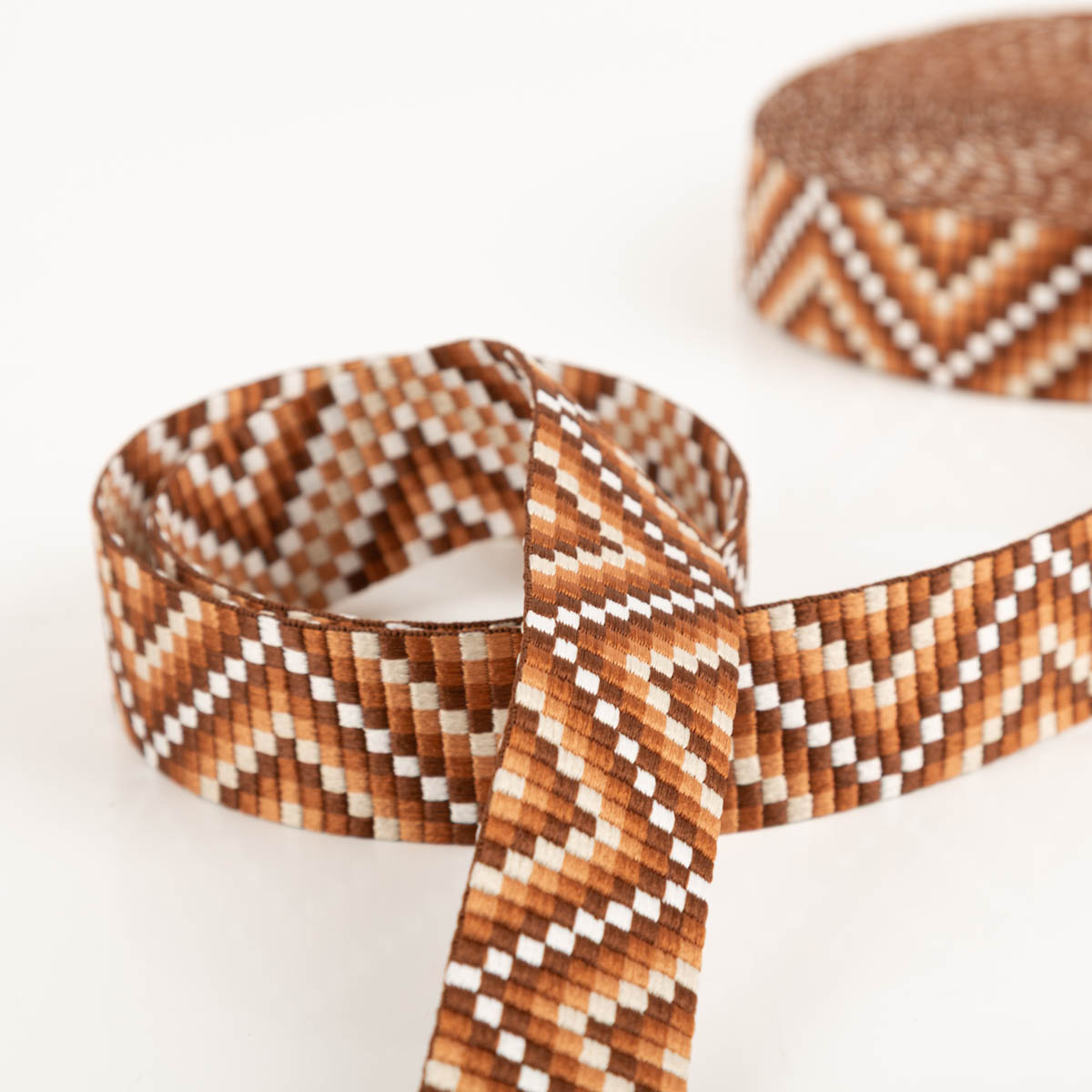 Gurtband Zickzack Chevron braun orange weiß 3,8cm Breite Breites Band mit Zickzackmuster in Braun, Orange und Weiß, zusammengerollt und teilweise entrollt auf weißem Untergrund